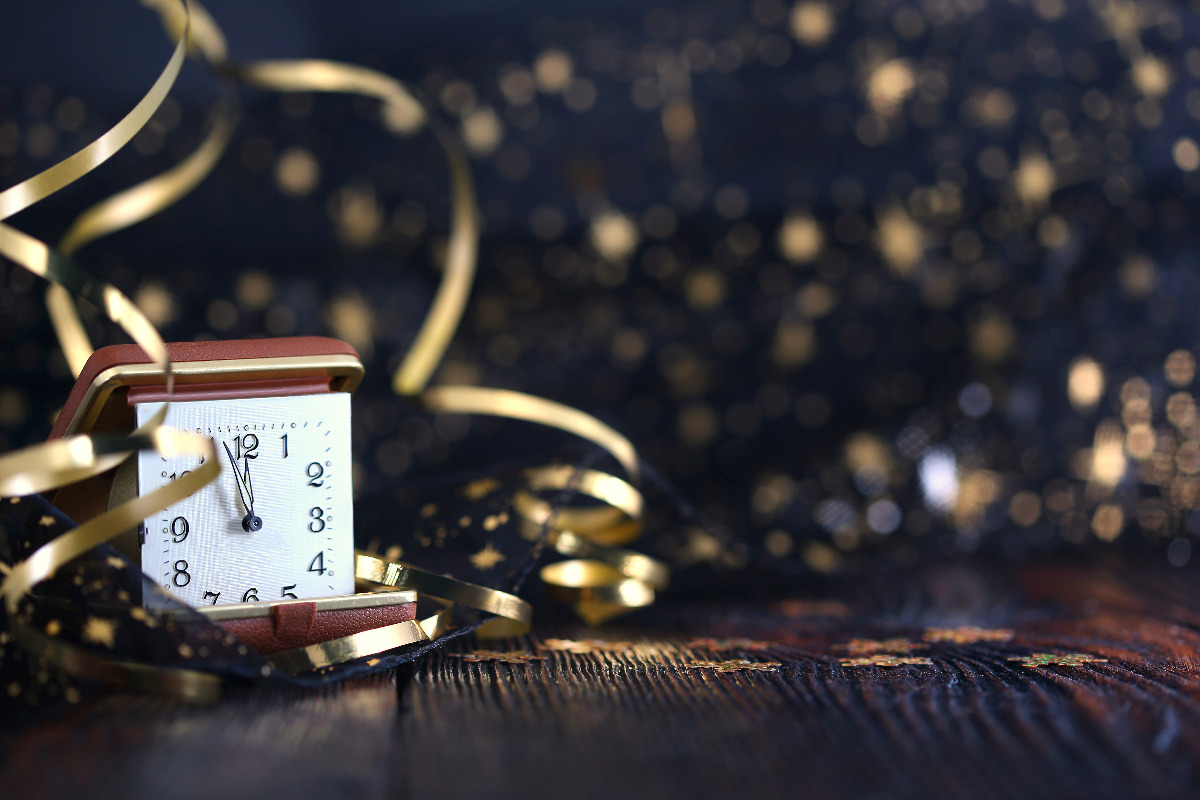 New Year's Eve - Corvallis, Oregon - A clock on a dark wood table showing nearly midnight, with gold glitter and confetti in the background & gold ribbons around the clock, against a dark backdrop